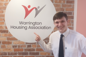 Young man with brown hair wearing a white shirt and dark tie standing in front on the Warrington Housing Association sign on a red brick wall in the WHA office